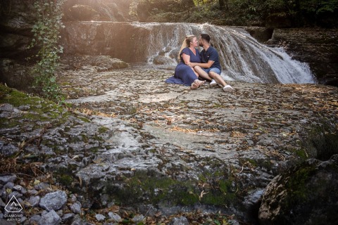 SASSENAGE, France — A couple sits bathed in sunlight below a cascading waterfall. This serene engagement portrait captures their quiet connection amidst a natural backdrop.