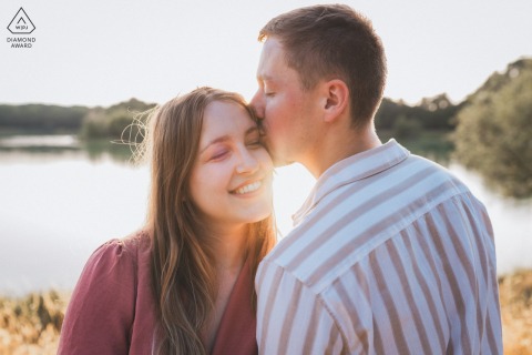 GIRONDE, France — A couple shares a tender kiss at sunset during their engagement portrait session at Lac Vert de Canéjan, a serene lake.