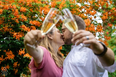 SEELEGER MOOR PARK, Zurich — A couple toasts their engagement with glasses of bubbly, sharing a kiss amidst a vibrant backdrop of blooming orange flowers in a stunning portrait.