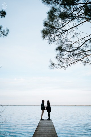 LACANAU, France — A couple stands at the end of a wooden dock, framed by tree branches and a blue sky, during their engagement portrait session on the serene shores of Lac de Lacanau.