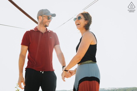 Laughing Couple Holding Hands In The Sunlight Against The White Wall LECTOURE, France — This engagement portrait captures a couple's playful connection as they laugh and hold hands in the sunlight against the bright white at The Castelnau.