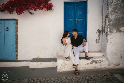 SANTORINI, Greece — A couple relaxes on a stoop of a classic white building with a vibrant blue door. This engagement portrait captures their quiet connection amidst the iconic streets of Santorini.