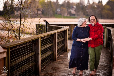 Love Walks Hand-in-Hand in the Rain at Rattray Marsh Engagement Portraits A couple walks hand-in-hand in the rain at Rattray Marsh, smiling lovingly, surrounded by lush, green, natural scenery.