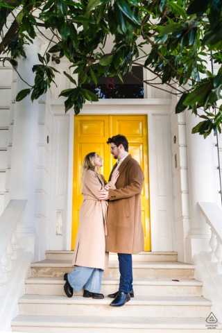 Engaged couple stands on staircase in London park, yellow door backdrop, smiling and embracing, modern romantic portrait.