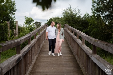 Engaged couple walks hand-in-hand on a rustic wooden bridge in Coubert, France, surrounded by lush greenery, smiling lovingly.