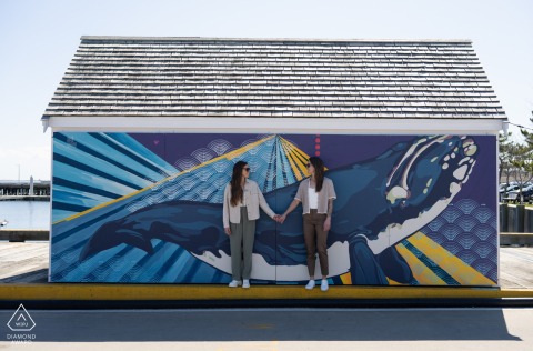 The couple stands in front of a vibrant whale mural in Provincetown, Massachusetts, lovingly glancing at each other, smiling.