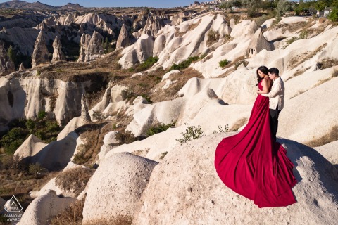 Bride-to-be in sweeping red dress stands elegantly with her future groom in Cappadocia desert, Goreme, Turkey, during pre-wedding formal photo session.