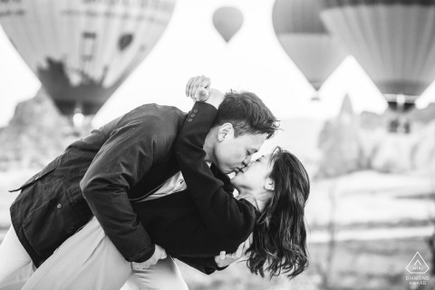 Black and white photo of couple dipping and kissing, hot air balloons floating in background, Cappadocia pre-wedding session, Goreme, Turkey.