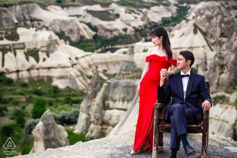 A formal couple poses in Cappadocia's desert: groom seated, bride standing beside him, both looking away.