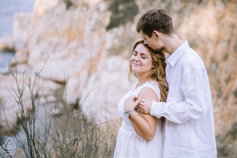 Tossa de Mar, Catalunya, Spain — A couple embraces on the cliffs as he holds her from behind, capturing a romantic and scenic portrait along the dramatic coastline.