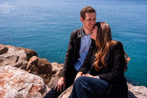 Portopiccolo, Sistiana, Trieste, Italy — A couple embraces by a sunlit sea stone wall, with water shimmering below them, capturing a romantic and scenic portrait by the coast.