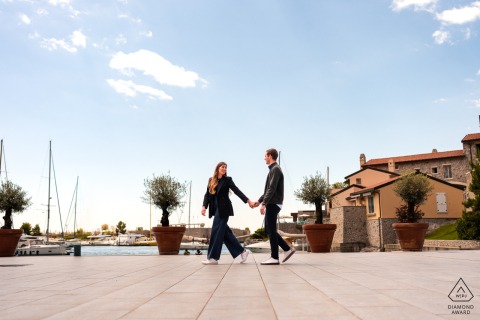 Portopiccolo, Sistiana, Trieste, Italy — A couple walks hand in hand by the waterfront as she leads and looks back at him, creating a playful and romantic portrait.