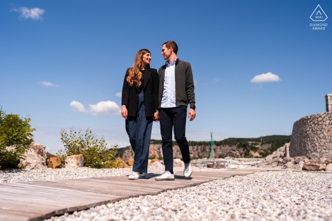 Portopiccolo, Sistiana, Trieste, Italy — A couple walks together on a sunlit wood boardwalk under a blue sky, captured from a low camera angle for a vibrant and modern portrait.