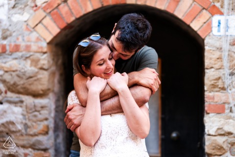 Grado, Gorizia, Italy — A couple shares a hug and kiss as he wraps her up from behind beside an Old World stone building, creating a warm and romantic portrait.