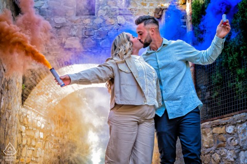 Spanish Romance: A Dramatic Smoke-Filled Kiss in the Old World Baeza, Jaén, Spain — A couple stands side by side, kissing and holding smoke grenades outside an Old World stone building, adding vibrant drama to this pre-wedding portrait.