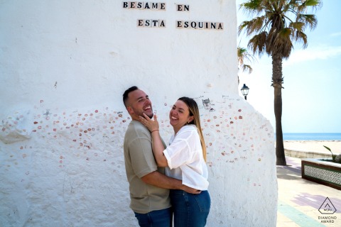 Rota, Cádiz, Spain — A couple stands facing each other and smiling beside a white stucco beach building, creating a bright and joyful pre-wedding portrait by the sea.
