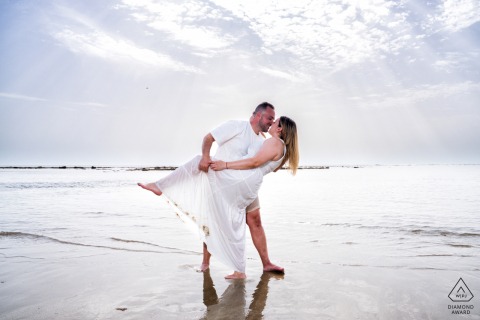 Rota, Cádiz, Spain — A couple, both dressed in white, dips at the ocean's edge during low tide, capturing a fresh and romantic pre-wedding portrait by the sea.