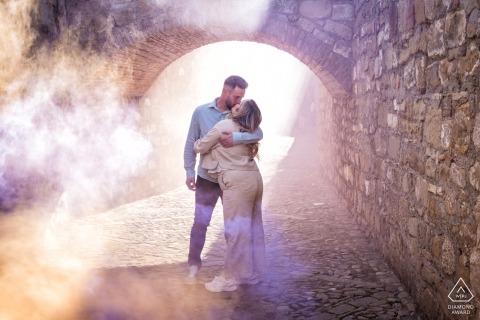 Baeza, Jaén, Spain — A couple embraces beneath an Old World stone arch as smoke drifts through the scene, creating a dramatic and romantic pre-wedding portrait.