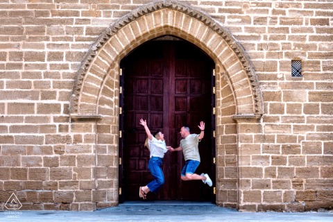 Rota, Cádiz, Spain — A couple leaps high together beneath the arch of an Old World stone building, capturing playful energy in this pre-wedding portrait.