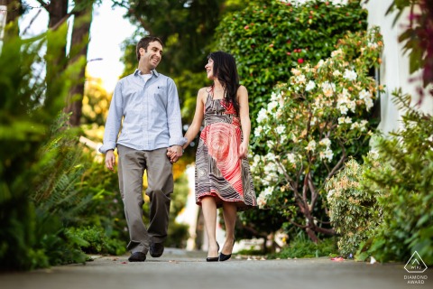 San Francisco, CA — A couple walks hand in hand, smiling along a paved path framed by lush foliage, captured from a low camera angle for a vibrant engagement portrait.