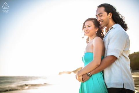 Santa Cruz, CA — He embraces her from behind as they share soft smiles on the beach near sunset, creating a warm and romantic engagement portrait.