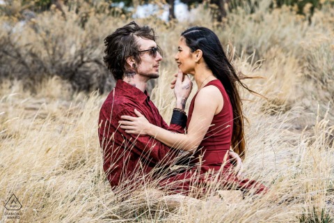 Denver, Colorado — A couple snuggles in tall grass under sunlight, dressed in matching burgundy, as the wind blows gently. This warm, natural engagement portrait highlights romance and unity in a stunning outdoor setting.