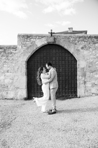 A couple poses for a romantic vertical black and white portrait in La Garde Adhémar, France, standing close together by an old stone building, framed by a doorway behind them.