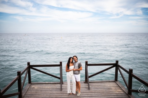 A newly engaged couple poses between the railings of Marbella Club Pier, symmetrically framed with water stretching out behind them, during their engagement photo session.