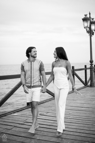 Marbella Pier Engagement: A Timeless Black and White Walk Hand in Hand A couple walks hand in hand along the Marbella Club Pier beside the water. The vertical black and white portrait captures their elegance and connection during their engagement photo session.