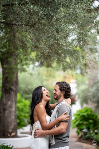 A newly engaged couple embraces and laughs together beneath the trees near the Marbella Club pier. The vertical portrait captures their joyful expressions during this engagement photo session.