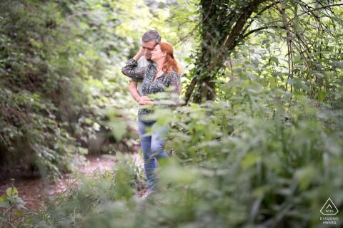 The engaged couple stands embracing near a tree and a small stream in a large park in Blagnac. They share an affectionate pose, surrounded by natural greenery during their engagement portrait session.
