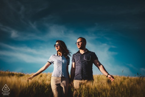 A couple walks closely together under a vivid blue sky in the sunlit, dry grass fields of Crete Senesi, Tuscany. The symmetrical framing highlights their connection in this countryside portrait.