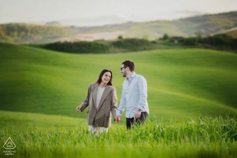A couple strolls together through the rolling hills of Crete Senesi, Tuscany, surrounded by a lush green landscape. The countryside’s gentle curves create a romantic backdrop for their engagement portrait.
