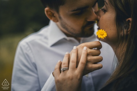 A couple stands in a field of flowers in Sorbolo Follo. He gently holds a single yellow flower near her face, both softly focused, creating a dreamy, romantic engagement portrait.