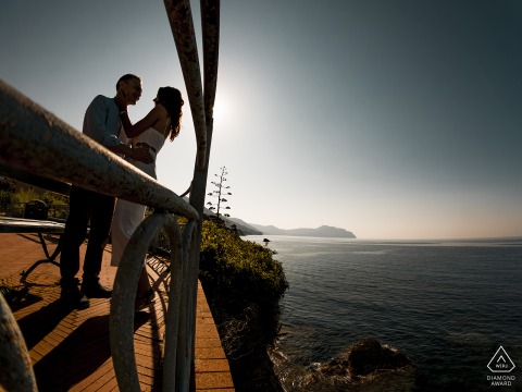 A couple stands at the railing of an overlook in Genova Nervi, their profiles silhouetted as they face each other. The dramatic lighting captures the romance of their engagement session.