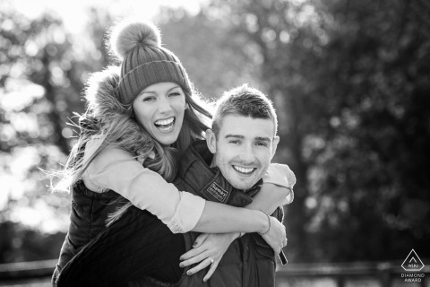 A playful black and white close-up of a couple at Lamport Manor Farm, with her riding piggyback on him. Both display big smiles, radiating fun and joyful energy during their engagement session.
