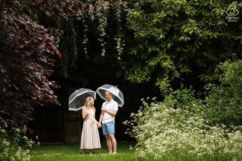 A couple stands together under a canopy of trees at Deene Park, each holding a clear umbrella. The lush foliage above creates a romantic backdrop for their engagement portrait session.