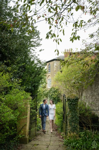 A couple walks together through a garden gate at Courteenhall Estate, surrounded by lush greenery. Their relaxed pose and the vibrant setting create a romantic engagement portrait.