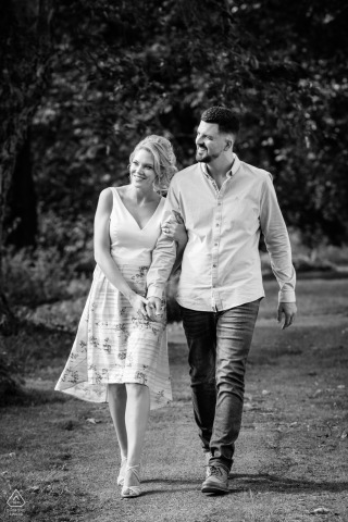 A couple walks hand in hand at Rushton Hall in a vertical black and white portrait. Both look off to the left, their expressions thoughtful, with elegant grounds as a backdrop.
