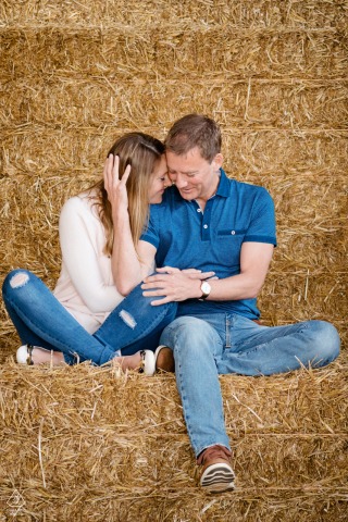 A couple sits closely snuggled together on a stack of hay bales at Lamport Manor Farm. Their relaxed pose and warm expressions capture a rustic, romantic atmosphere during their engagement session.