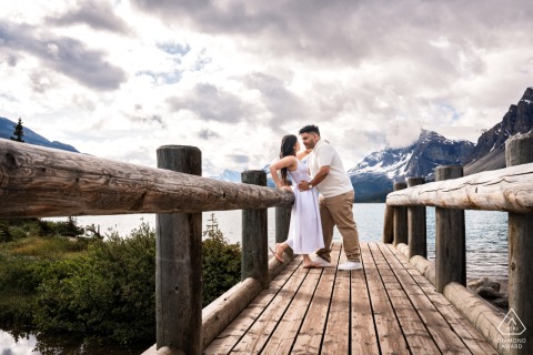 An engaged couple shares a warm embrace on the bridge at Bow Lake, Canada. He holds her gently, her back resting against the wooden path railing, surrounded by the tranquil natural landscape.