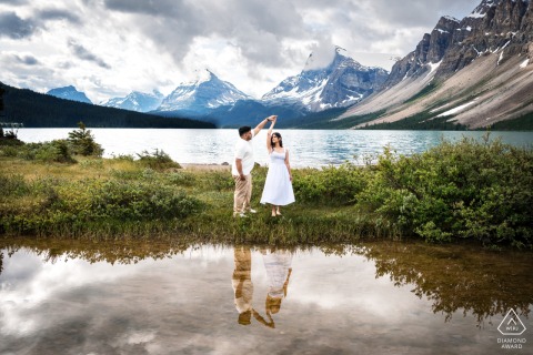 An engaged couple dances by the edge of Bow Lake, Canada, with the water below them reflecting surrounding mountains and clouds. The serene setting enhances the romance of their engagement portrait.