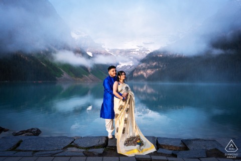 A man embraces his fiancée at Lake Louise, Canada, standing close together with bright, shimmering water and low clouds in the background, creating a serene and romantic portrait.