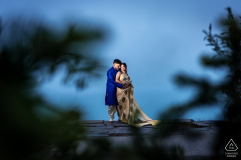 A couple embraces lovingly at Lake Louise, Canada, captured through softly focused bushes. Green foliage frames the image on each side, while a vibrant blue sky completes the romantic engagement portrait.