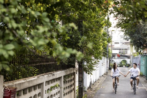Bangkok alley engagement photo of a couple riding bikes, holding hands, and smiling at each other.