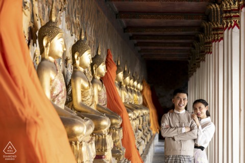 A couple poses for their engagement portrait in Bangkok, Thailand, standing beside a row of ornate old world gold statues on the left, which adds a touch of cultural elegance to the composition.
