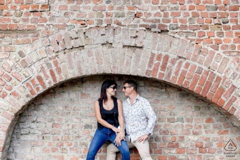 In Saint Pierre de Bressieux, France, a couple sits beneath a red brick arch on a historic building, gazing affectionately at each other during their engagement portrait session.