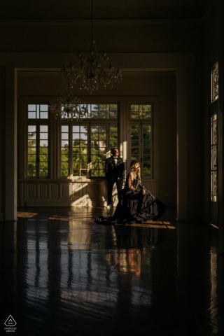 In São Paulo, a stylish couple poses indoors, illuminated by harsh dramatic sunlight streaming through large windows. The bold lighting and modern setting create a striking and contemporary engagement portrait.