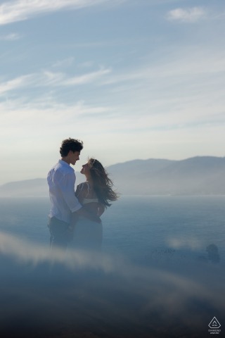 At the top of Point Dume Natural Preserve in Malibu, California, a couple poses with sweeping views of the beach below. Misty, dreamlike light and a blue sky add a romantic atmosphere to their engagement portrait.