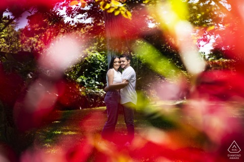 At Hotel Bougainvillea in Santo Domingo, Heredia, Costa Rica, a couple is romantically framed by vibrant red flowers in the foreground, set against the lush backdrop of Bougainvillea Gardens during their engagement portrait session.